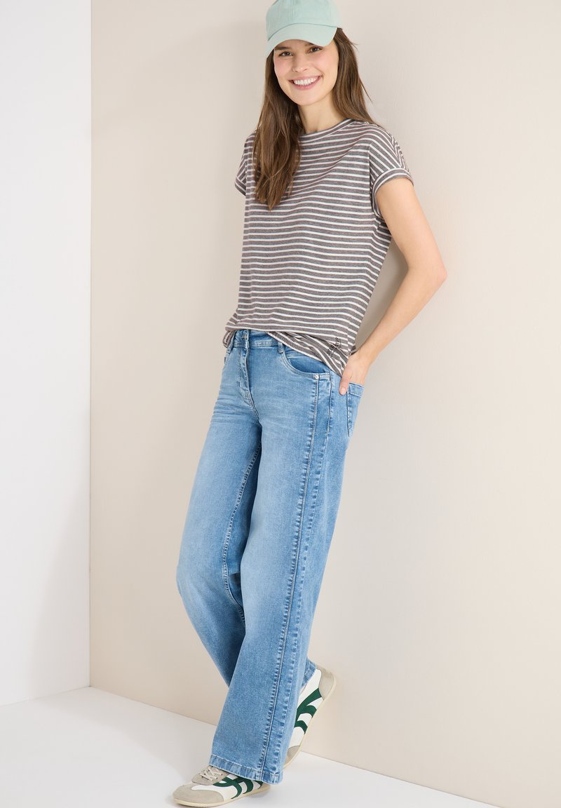 Jeune femme souriante, portant un t-shirt rayé, un jean bleu clair, des baskets et une casquette vert clair, debout devant un mur beige.