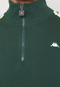Sweat-shirt zippé vert avec col haut, présentant une fermeture éclair texturée et un logo contrasté sur la poitrine. Tissu lisse et design athlétique.