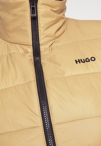 Světlé béžové péřové bundy s vysokým límcem, černým zipem vpředu a výrazným černým logem "HUGO" na hrudi. Hladká textura.