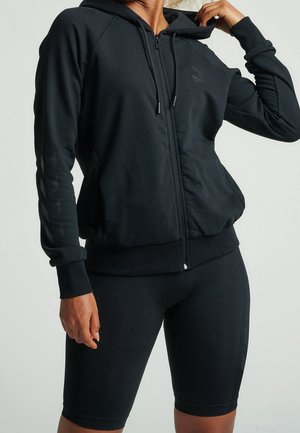 Person trägt schwarzen Kapuzenpullover mit Reißverschluss und Kordelzug sowie schwarze enganliegende Radlerhosen und steht vor einem schlichten Hintergrund.