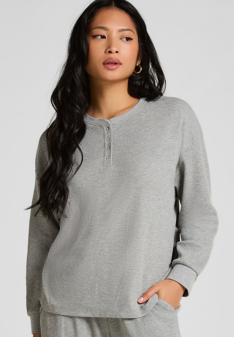 Grå långärmad henley-tröja i texturerad våffelstickning; har rund hals, fyraknappslå och avslappnad passform med lätt rundad nederkant.