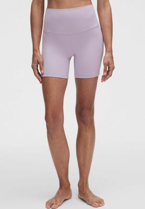 Femme portant un short de sport ajusté violet clair taille haute, debout pieds nus sur un fond clair uni.