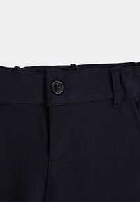 Ceinture de pantalon bleu marine foncé avec un bouton noir à quatre trous, passants de ceinture et coutures visibles sur un fond clair uni.
