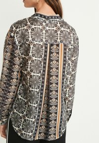 Schwarz-weiß gemusterte Bluse mit floralen Designs, ausgestattet mit einem Kragen und langen Ärmeln, aus leichtem Stoff. Akzente in Beige und Orange.