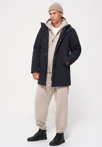 Ciemnoniebieska parka z kapturem, kontrastujące beżowe wewnętrzne warstwy, zamek i zatrzaski, oraz boczne kieszenie, w zestawieniu z beżowymi spodniami dresowymi i czarnymi butami.