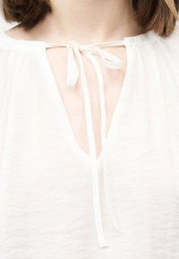 Λευκό πουκάμισο με λαιμόκοψη σε σχήμα κλειδαρότρυπας, δεμένο με λεπτές υφασμάτινες λωρίδες, που αποκαλύπτει την περιοχή της κλείδας και του λαιμού ενός ατόμου.