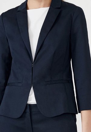 Kobieta ubrana w dopasowaną granatową marynarkę z przednimi kieszeniami na białą bluzkę, w połączeniu z pasującymi granatowymi spodniami.