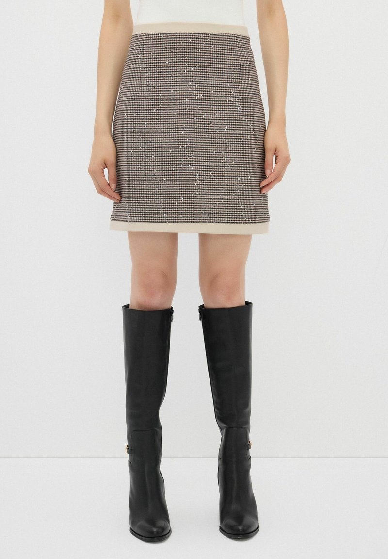 Jupe à motif pied-de-poule avec une taille beige, longueur genoux, complétée par des bottes noires à hauteur de genou avec une finition lisse et une fermeture éclair.