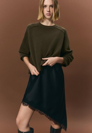 Olíva zöld kötött pulóver fekete, aszimmetrikus, csipkedíszes szoknyával párosítva. Puha textúra, kényelmes szabás és kifinomult részletek.