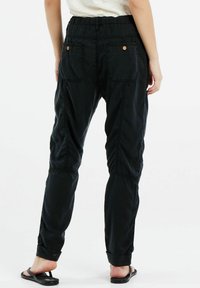 Zwarte stoffen broek met een elastische tailleband, twee achterzakken en opgerolde mouwen. Gelaagd oppervlak met zichtbare stiksels.