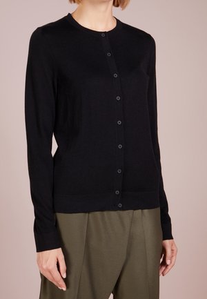 Woman wearing a black button-up cardigan with long sleeves and olive green pants, standing against a plain beige background.