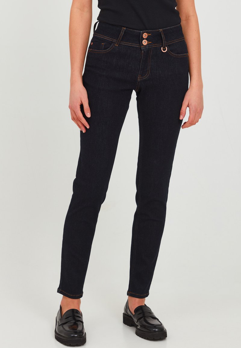 Jeans skinny de mezclilla azul oscuro con cintura alta, con detalles de botones de cobre y costuras naranjas. Textura suave, ajuste entallado y longitud hasta los tobillos.