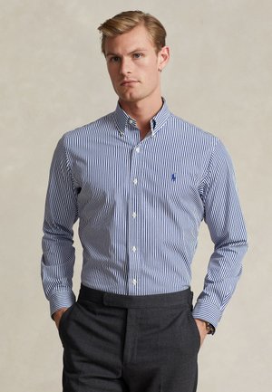 Mann, der ein blau-weiß gestreiftes Hemd mit Knöpfen und dunkelgraue Hose trägt, steht mit den Händen in den Taschen vor schlichtem Hintergrund.