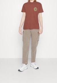 Brun bomullströja med grön blommönster på vänster bröst, kombinerad med beige cargobyxor och vita sportskor.