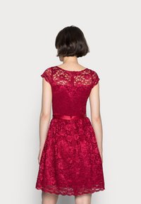 Robe rouge en dentelle avec manches courtes style cape, corsage ajusté et jupe évasée. Caractérisée par une ceinture en satin et un motif floral sur l'ensemble.