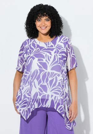 Uśmiechnięta kobieta z kręconymi czarnymi włosami, ubrana w luźną fioletowo-białą bluzkę w liście i jednolite fioletowe spodnie na jednolitym tle.