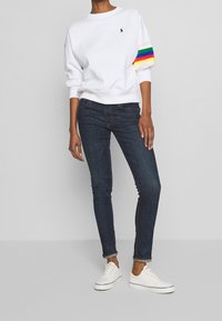 Bílá mikina s vícero barevnými pruhy na rukávech, tmavě modré úzké džíny a bílé tenisky s nízkým profilem a tkaničkami.