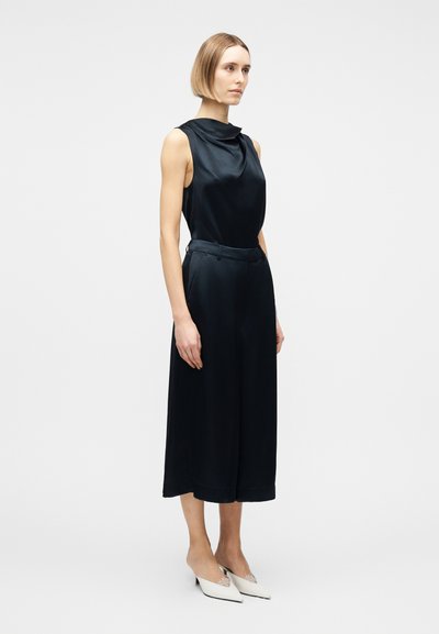 Γυναίκα με κοντά ξανθά μαλλιά που φοράει αμάνικο μαύρο σατέν τοπ, παντελόνι με φαρδιά μπατζάκια σε ίδιο χρώμα και λευκά μυτερά mule με τακούνι.