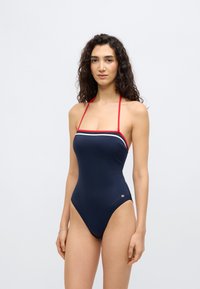 Σκούρο μπλε ολόσωμ ο μαγιό με κόκκινες και λευκές ρίγες; κατασκευασμένο από ελαστικό υλικό; διαθέτει λουριά γύρω από το λαιμό και σχεδιασμό με ψηλότερο κόψιμο στα πόδια.