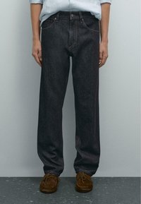 Modellen står foran en ensfarvet baggrund iført mørke denim jeans og brune sko, med hænderne afslappet ned langs siden.