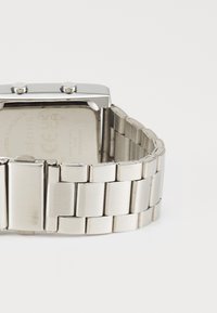 Montre-bracelet en métal argenté avec un cadran rectangulaire, deux boutons poussoirs et un bracelet en maillons brossé. Le dos présente des détails gravés.