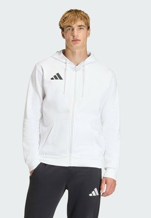 Jeune homme aux cheveux blonds portant un sweat à capuche blanc zippé et un pantalon de survêtement noir, tous deux avec un logo triple bandes noir et blanc.