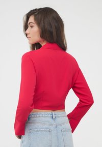 Maglietta rossa cropped con colletto, maniche lunghe e una texture liscia. Indossata sopra pantaloni corti di jeans a vita alta, che mettono in risalto una silhouette aderente.