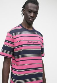 T-shirt a righe in rosa, blu navy e crema. Presenta scollo a coste e ricamo "Guess Originals" sul petto. Maniche corte, tessuto di cotone.