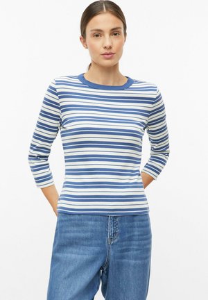 Frau mit blau-weiß gestreiftem Langarmshirt und blauen Jeans, steht mit den Händen hinter dem Rücken vor einem einfarbigen Hintergrund.