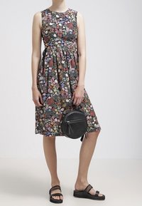 Ärmelloses Kleid mit floralem Muster aus dunklem Stoff und gerafftem Bund. Kombiniert mit einer schwarzen runden Handtasche und schwarzen Plateausandalen.