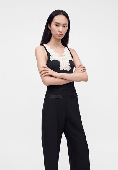 Femme aux longs cheveux noirs portant un top noir sans manches avec un détail en dentelle blanche et un pantalon noir, debout les bras croisés devant un fond blanc.