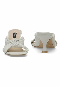 White leather slide sandals feature a knotted design on the upper and a modest, curved heel. The sole is beige and smooth.