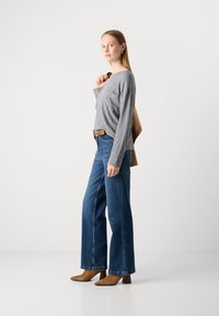 Kvinne stående i grå langermet topp, vide blå jeans, brune helte støvler, som holder en brun jakke over skulderen mot en hvit bakgrunn.