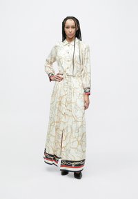 Maxi kleita baltā audumā ar zelta ķēdes rakstu, ar pogām priekšpusē, garām piedurknēm un tumši zilu/sarkanu svītrainu apmali. Viduklis ar jostas detaļu.