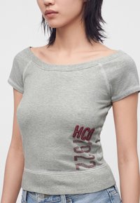 Woman wearing gray off-shoulder short-sleeve top with red "HCO 1922" text and light blue jeans.