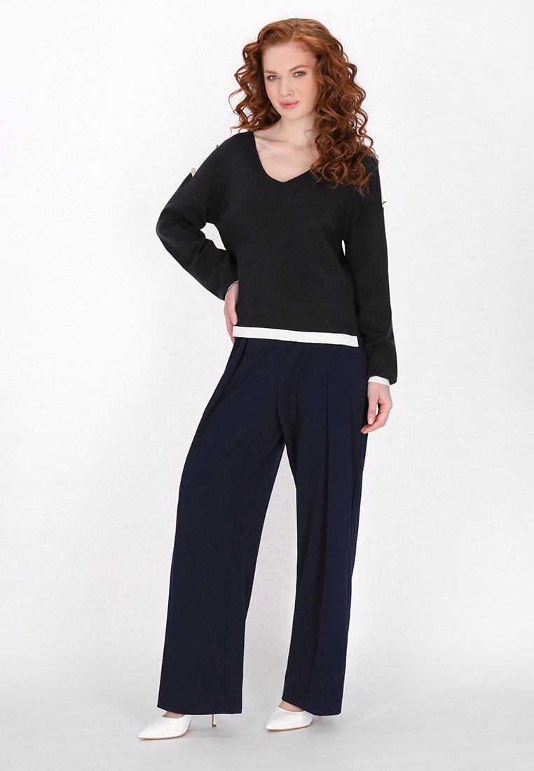 Czarny sweter z dekoltem w serek i białym wykończeniem, zestawiony z granatowymi szerokimi spodniami. Modelka stoi w białych szpilkach, prezentując swobodny strój.