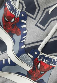 Felpa grigia con un grafico di Spider-Man su scarpe da ginnastica blu, con lacci bianchi e dettagli di cucitura. Il testo recita "SPIDER".