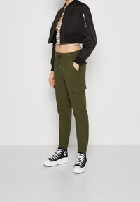 Svart kort bomberjacka med zipficka, matchad med olivgröna cargobyxor med justerbar midja och sidofickor. Svarta högställda sneakers.