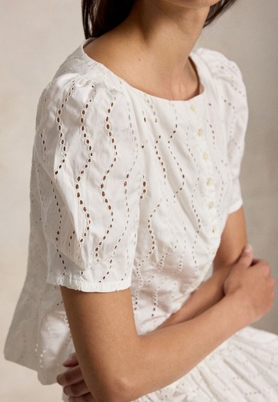 Γυναίκα που φοράει λευκό μπλουζάκι με puff μανίκια και κουμπιά στο μπροστινό μέρος, με σταυρωμένα τα χέρια, μπροστά σε ουδέτερο φόντο.