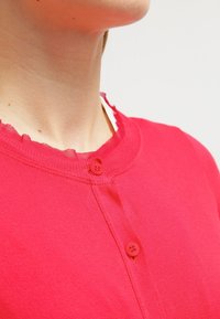 Cardigan en coton rouge avec col rond, présentant une finition brute au niveau du col et une fermeture à un bouton. Texture lisse avec une couleur unie.