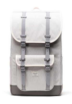 Sac à dos en tissu matelassé gris avec fermeture à rabat, doté de bretelles grises et d'une bordure blanche. Comprend une étiquette logo à l'avant et un fond renforcé.