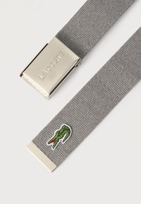 Ceinture en tissu gris tissé avec boucle argentée embossée du mot "LACOSTE" et logo crocodile vert brodé près de l'extrémité.