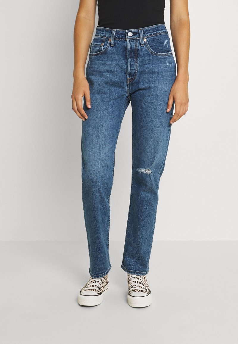 Jeans bleu taille haute en denim, avec une coupe droite, une légère usure au genou droit et des détails de surpiqûres discrètes.