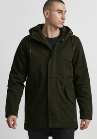 Parka olive verte avec capuche, dotée d'une fermeture éclair à l'avant, de deux poches latérales et d'une coupe légèrement ample. Fabriquée en tissu épais.