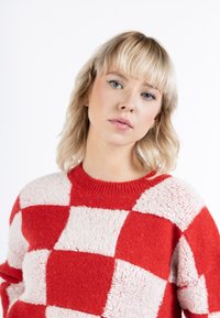 Suéter de patrón a cuadros en rojo y crema, hecho de un material de punto esponjoso, con un cuello redondo acanalado y mangas largas.