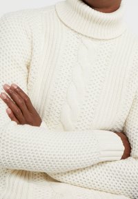 Maglione lavorato a maglia di colore crema con colletto alto. Presenta motivi testurizzati, trecce e polsini a costine. Vestibilità morbida con lavorazione grossa.