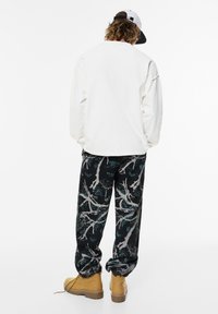 Chemise blanche à manches longues, pantalon noir avec un motif de branches d'arbre et bottes jaunes. Présente une touche de bleu foncé et une coupe décontractée.