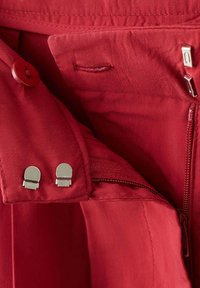 Ceinture en tissu rouge avec bouton, fermeture éclair et double agrafe métallique sur un vêtement.