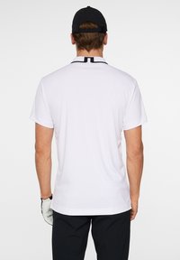 Vit golfpolo med korta ärmar och en svart detaljer vid kragen, tillverkad av ett lätt tyg med en slät textur.