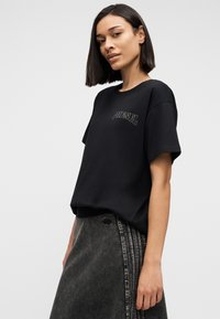 Γυναίκα που φοράει μαύρο T-shirt με το λογότυπο Diesel και σκουρόχρωμη γκρι φούστα τζιν με ψηλή μέση, στέκεται απέναντι σε ένα απλό ανοιχτό φόντο.